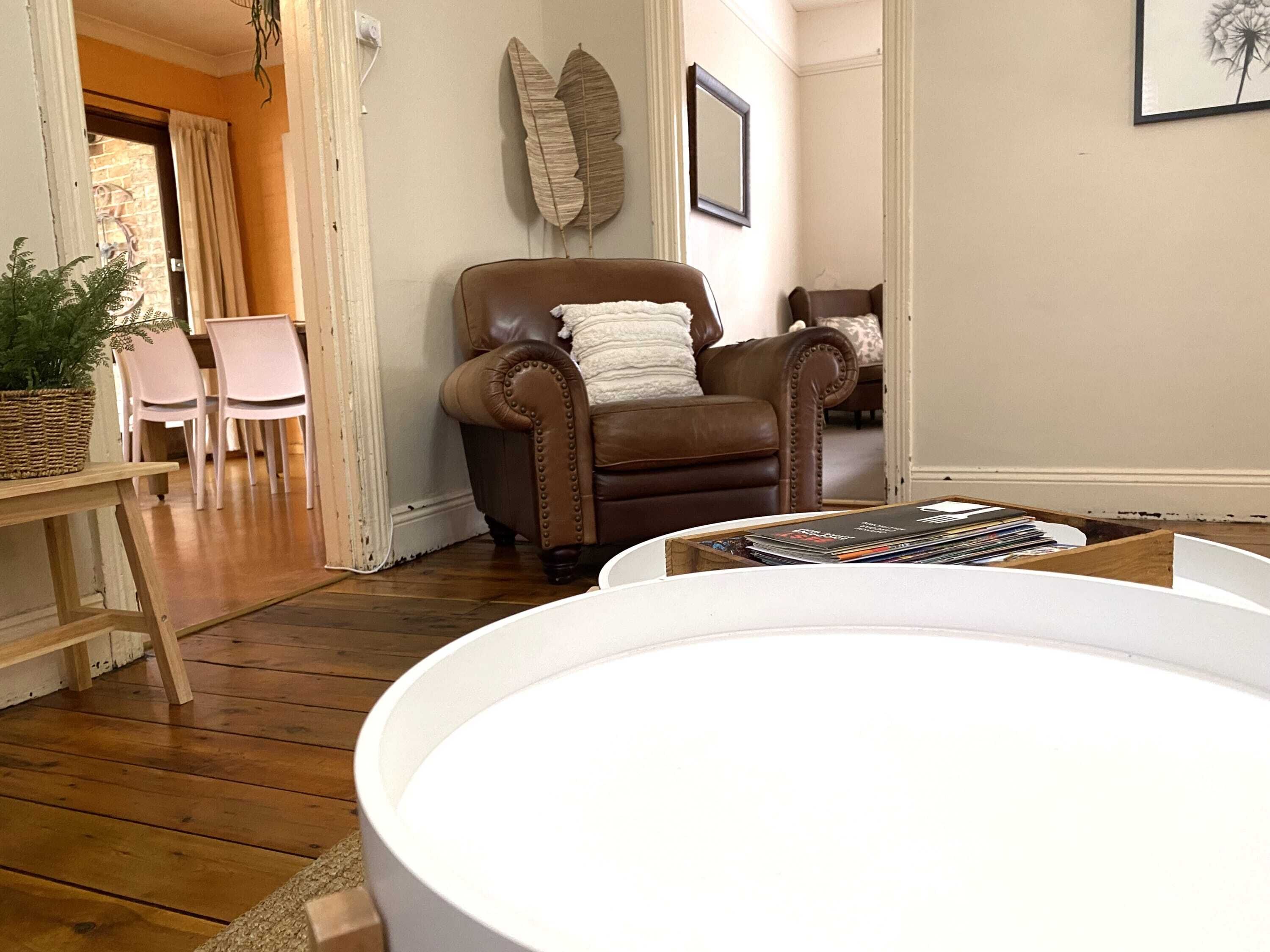 A living room with a brown leather armchair, a white armchair, a dining table with chairs, and a bookshelf.