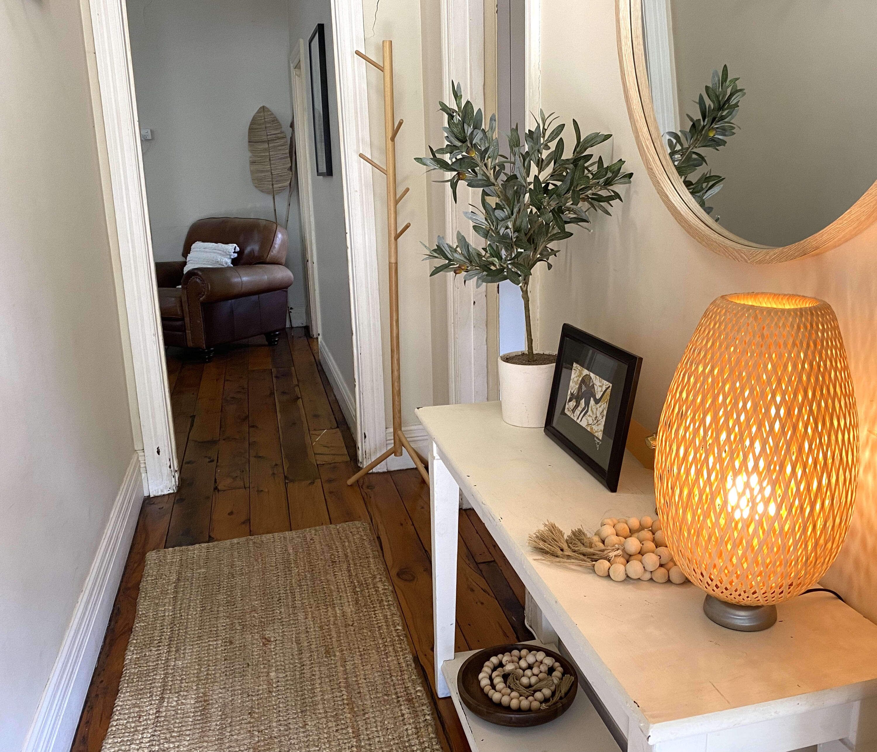 Un couloir doté d’un plancher en bois, d’un canapé en cuir marron, d’une plante en pot, d’un tableau encadré, d’une lampe en osier et d’un bol rempli d’objets décoratifs.