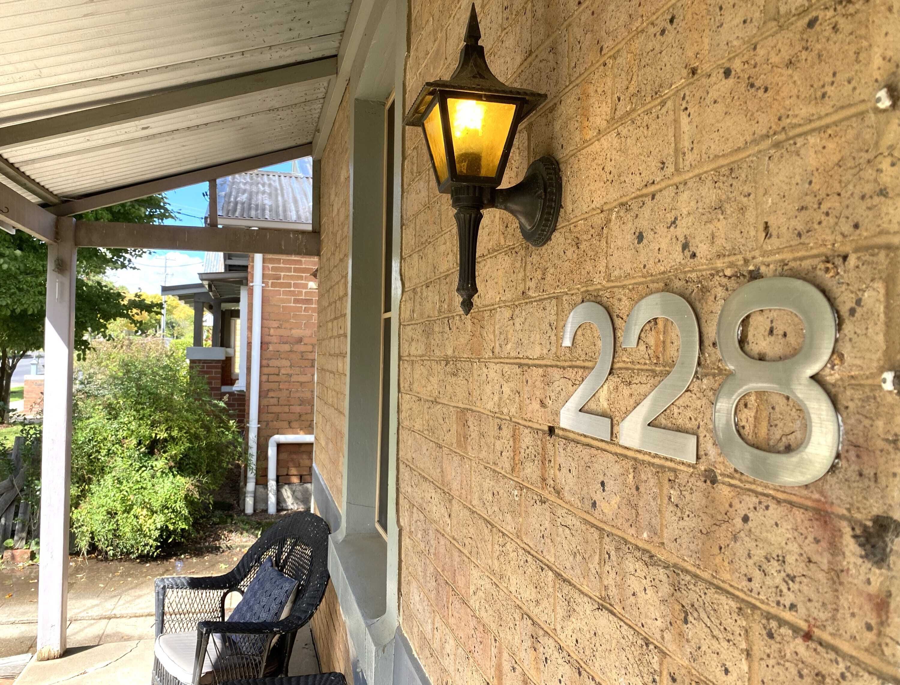 A house number 228 on a brick wall, a black outdoor chair, and a wall-mounted lamp.
