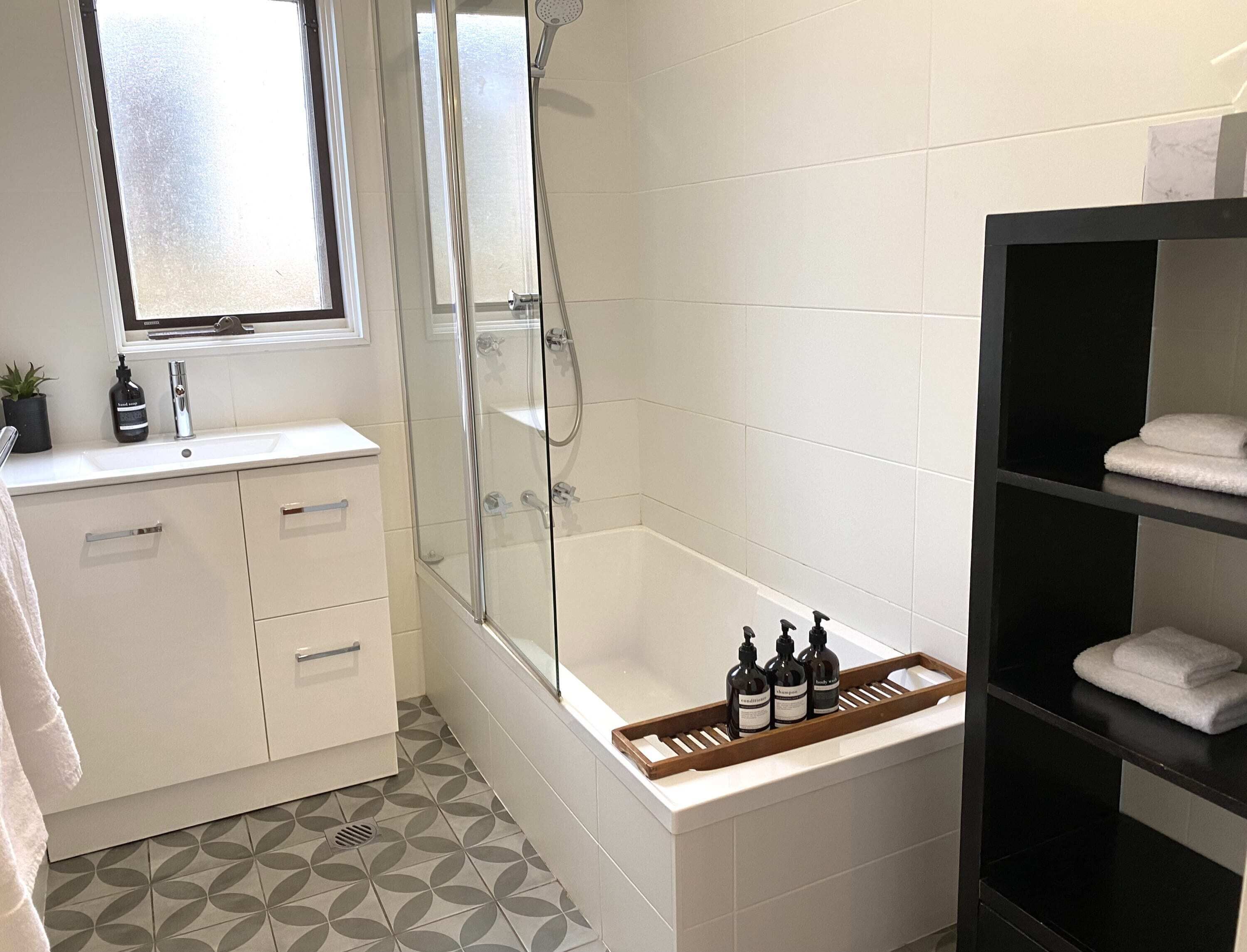 A modern bathroom with a glass-enclosed shower, a white sink with a mirror, and a wooden shelf holding toiletries.