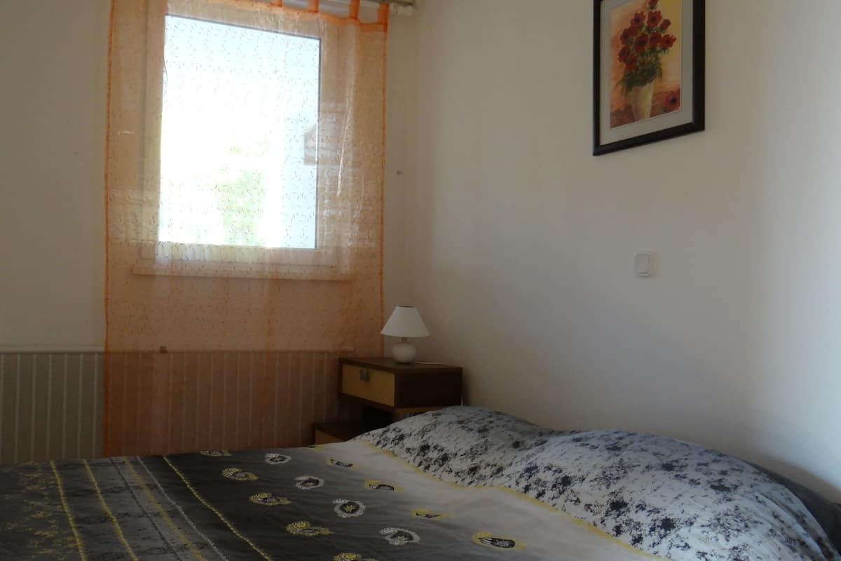 A bedroom with a bed, a window with sheer curtains, a bedside table with a lamp, and a framed picture on the wall.