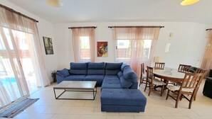 A living room with a blue sectional sofa, a dining table with wooden chairs, and large windows with sheer curtains.