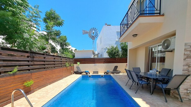 A swimming pool with a wooden deck, lounge chairs, and a table with chairs.