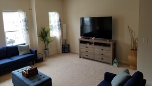 Un salon avec un grand téléviseur à écran plat posé sur un meuble en bois, un canapé bleu, un pouf bleu, un ventilateur de plafond et une plante en pot.