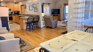 A modern kitchen with wooden cabinets, a dining area with a marble table, and a living room with a grey armchair and a TV.