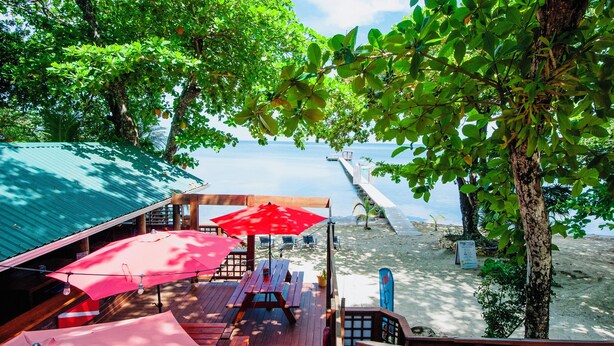 Una terraza de madera con mesas de picnic y sombrillas rojas con vista a una playa.