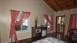 Amplia habitación con techo alto, vigas de madera, ventilador de techo, gran ventana con cortinas rosadas, una pequeña mesa con una planta y vistas al exterior.