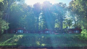 A row of cabins by a lake with a grassy embankment and dense trees in the background. - Torch Lake Cabin Rentals~ Where Cozy Lakefront Cabins meet stunning Torch Lake! (Kewadin)
