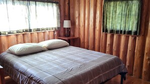 A wooden room with a bed, a nightstand, a lamp, and a window with curtains. - Torch Lake Cabin Rentals~ Where Cozy Lakefront Cabins meet stunning Torch Lake! (Kewadin)