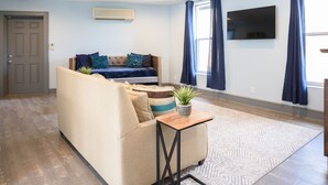 Ett vardagsrum med en beige soffa, ett litet träbord med en växt, en mönstrad matta, en tv på väggen och en dörr i bakgrunden.