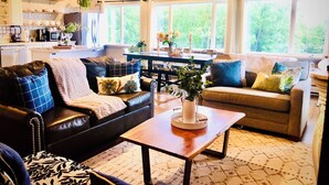 A cozy living room with a black leather sofa, a wooden coffee table, and a beige sofa with blue cushions. A dining area with a table and chairs is visible in the background. - Lovely 3br/2ba Hideaway! Great for Groups & Families! Inlet & Denali Views (Anchorage)