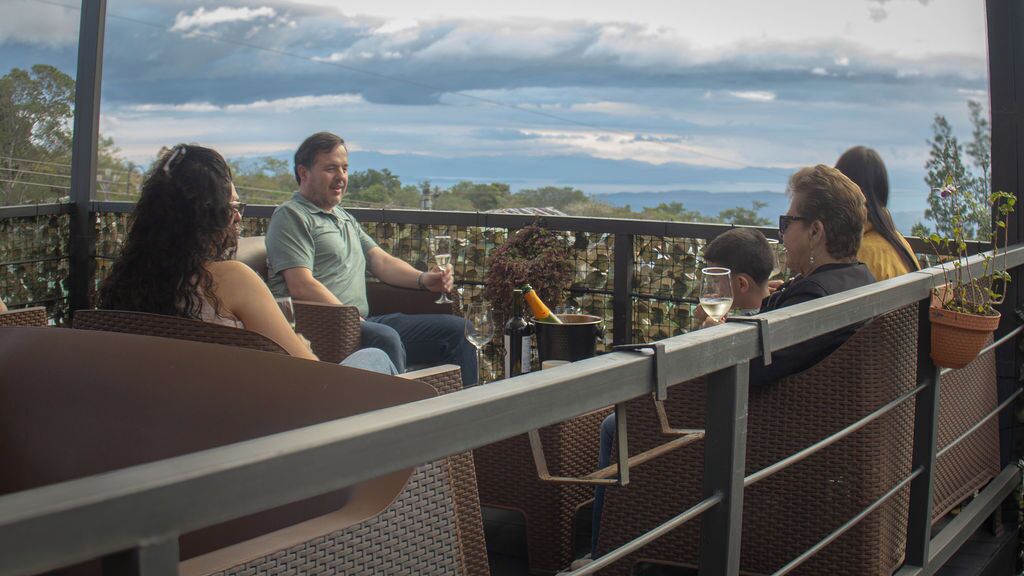 Un groupe de personnes est assis à la terrasse d’un café, entouré de meubles en osier, et profite de la vue sur le paysage.
