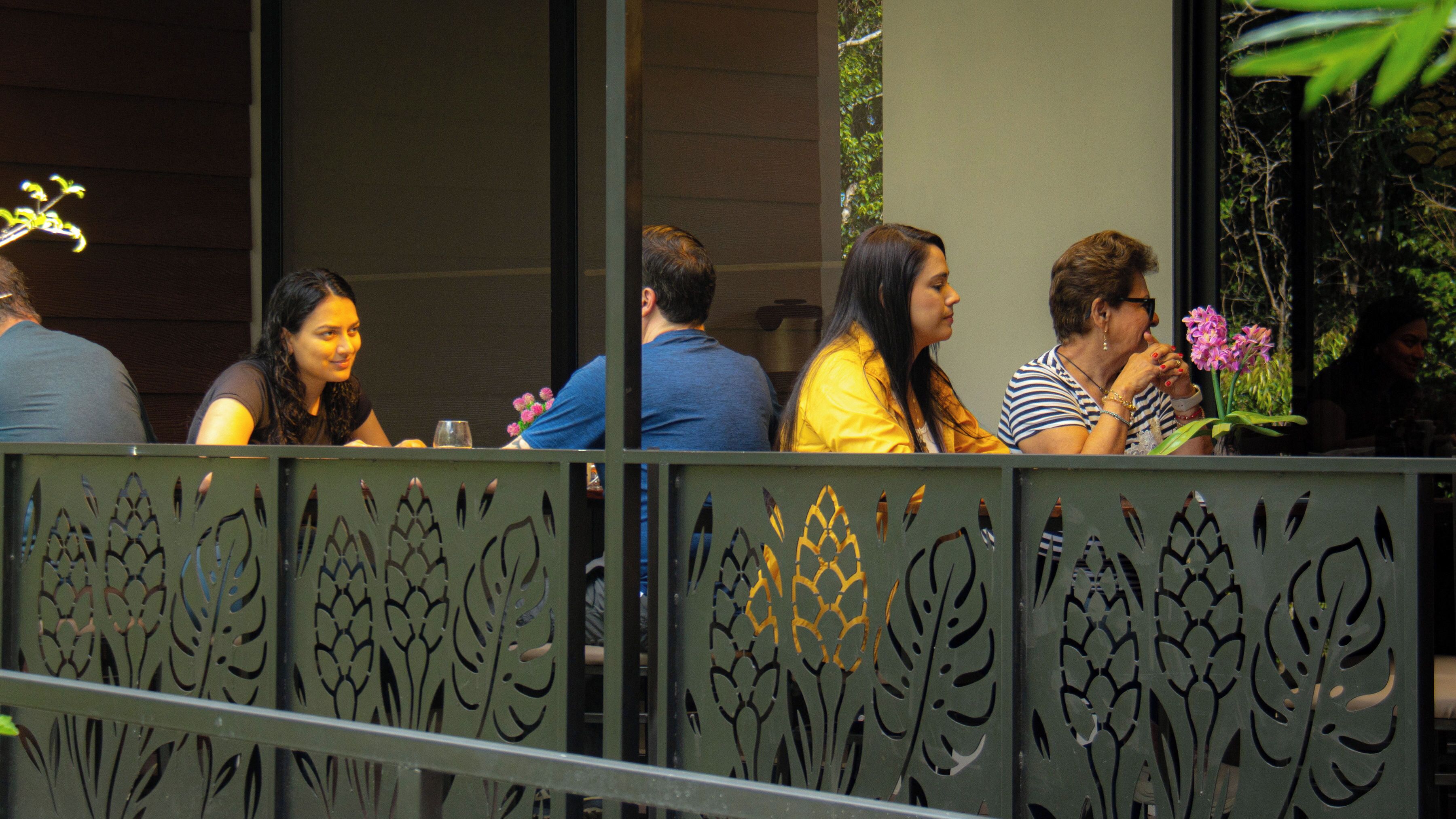 Un groupe de personnes est assis à une table dans un espace de restauration en plein air, lequel est agrémenté d’une rambarde en métal décorative.