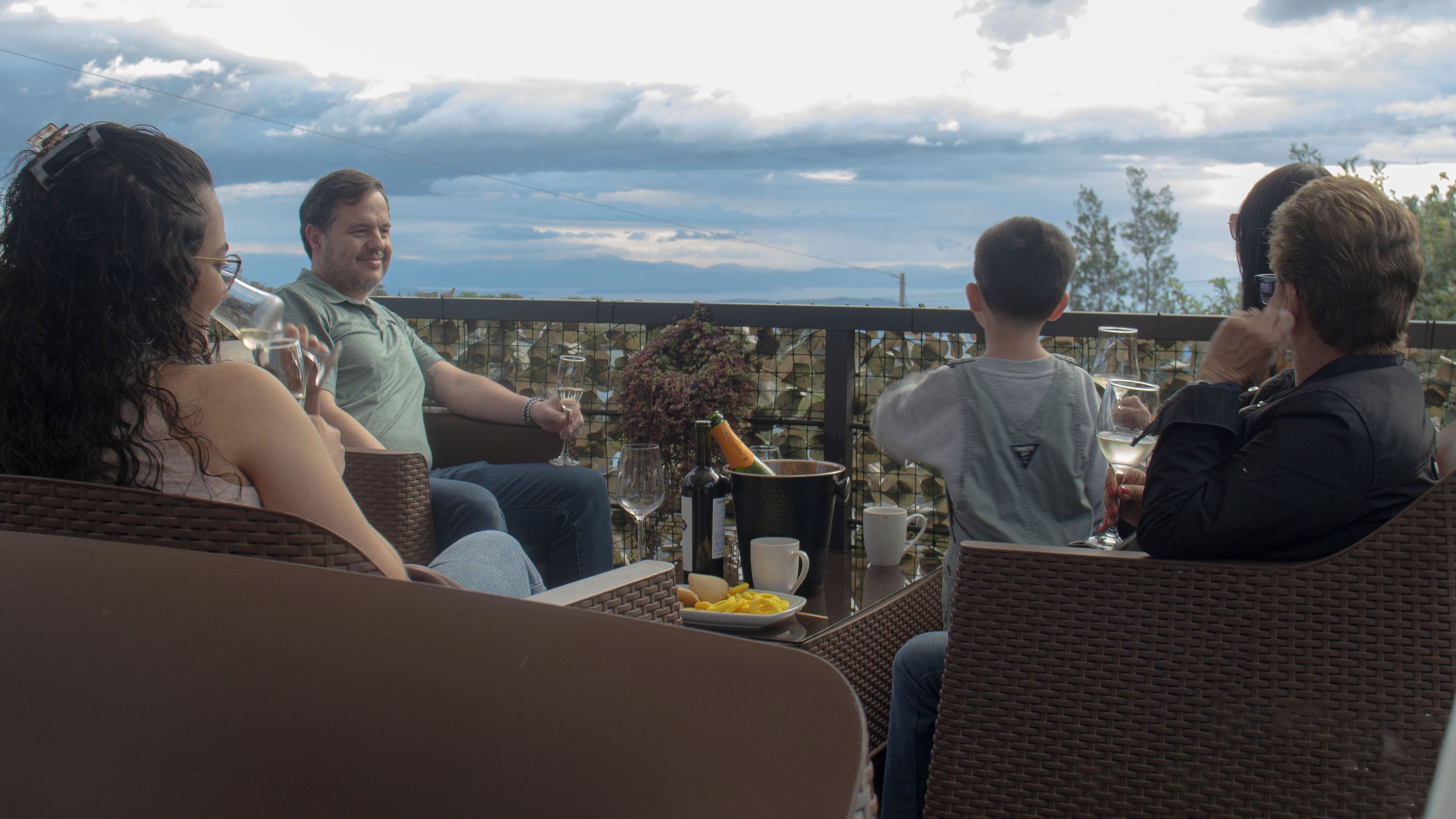 A group of people seated on wicker chairs at an outdoor table with a bottle of champagne and a plate of food.