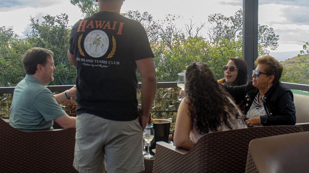 A group of people seated at an outdoor table with wicker furniture, a man standing in the background wearing a black t-shirt with 'HAWAII' and 'BIG ISLAND TENNIS CLUB' on it.