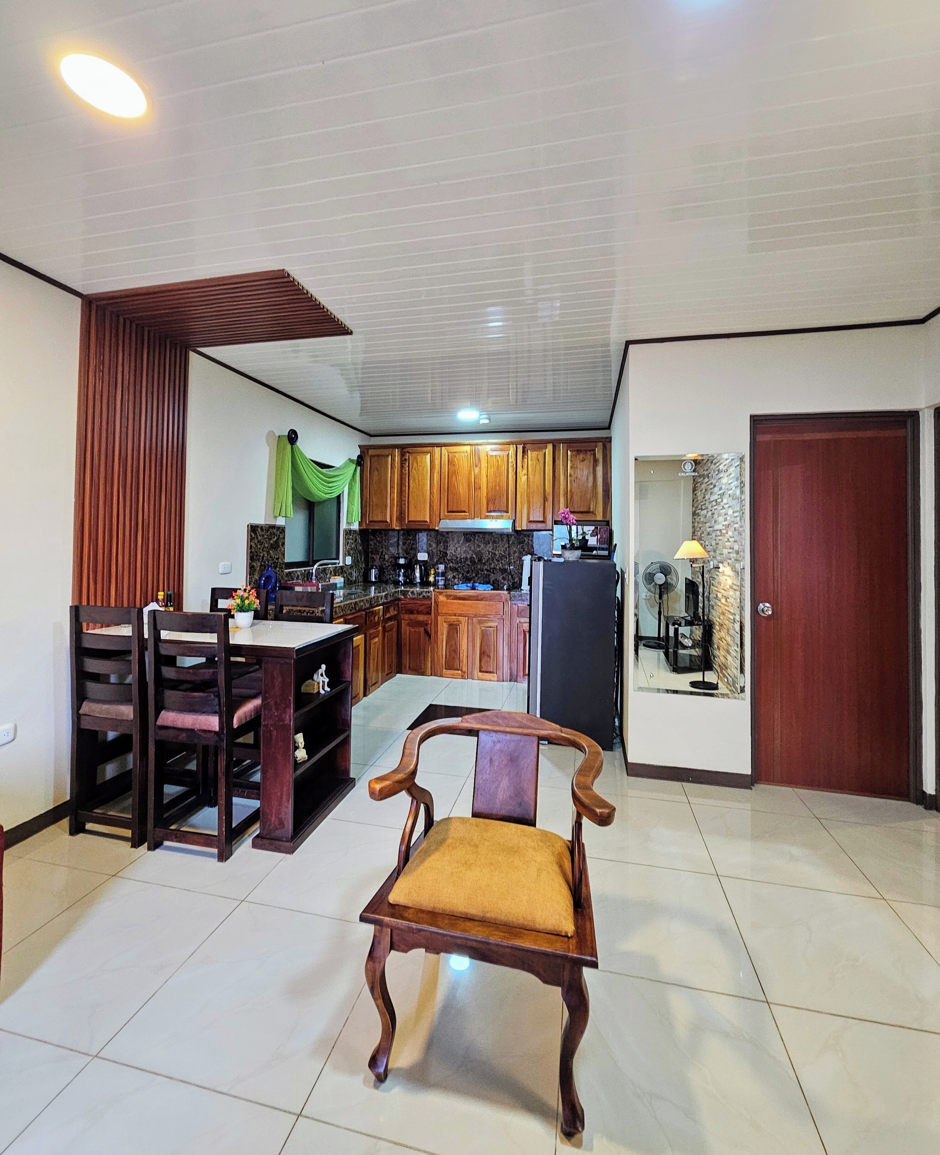 A spacious living area with a wooden dining table, a wooden chair with a cushion, and a kitchen in the background with wooden cabinets and a refrigerator.