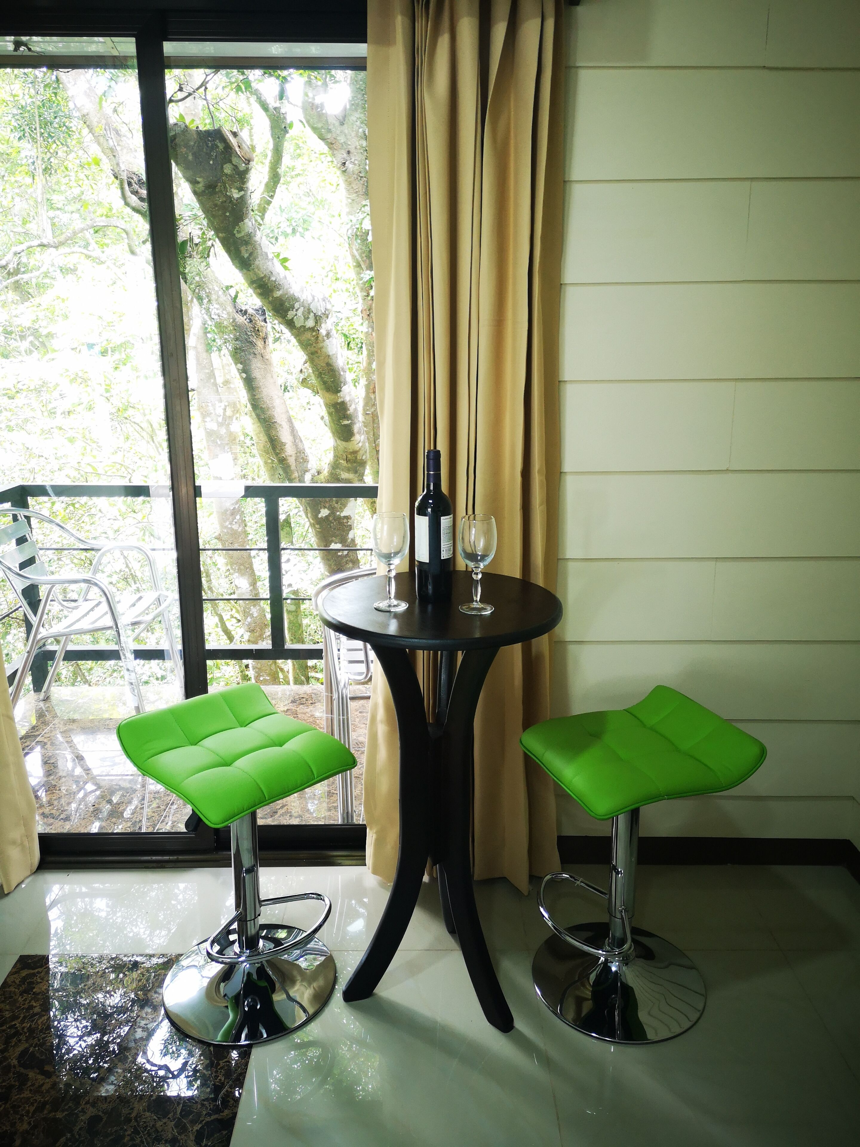A table with two green stools, a bottle, and two wine glasses.