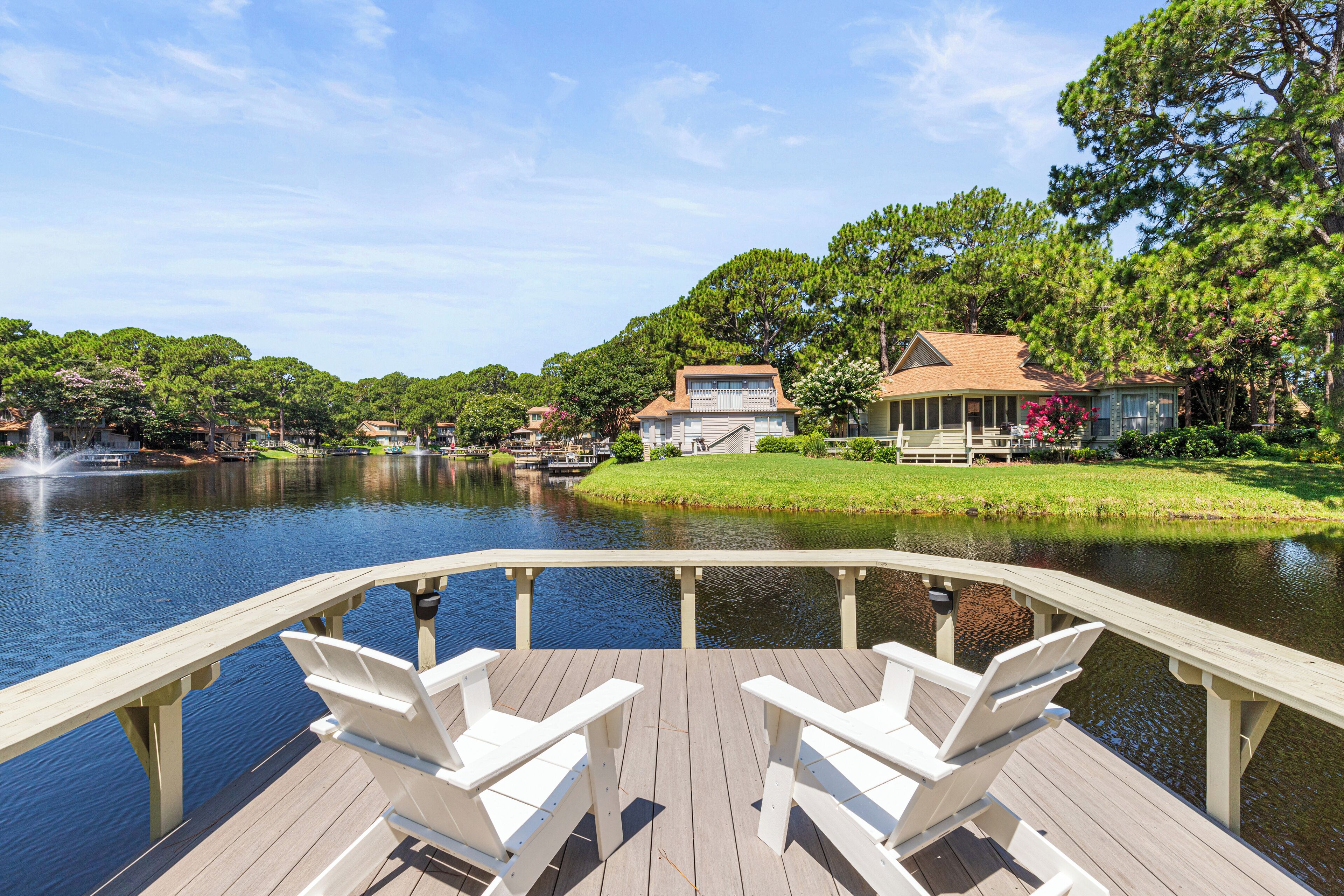 Eine Holzterrasse mit weißen Stühlen, die auf einen See mit umliegenden Bäumen und einem Haus blickt.