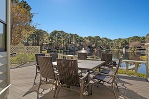 A deck with wicker chairs and a table overlooking a lake with boats, surrounded by trees and houses.