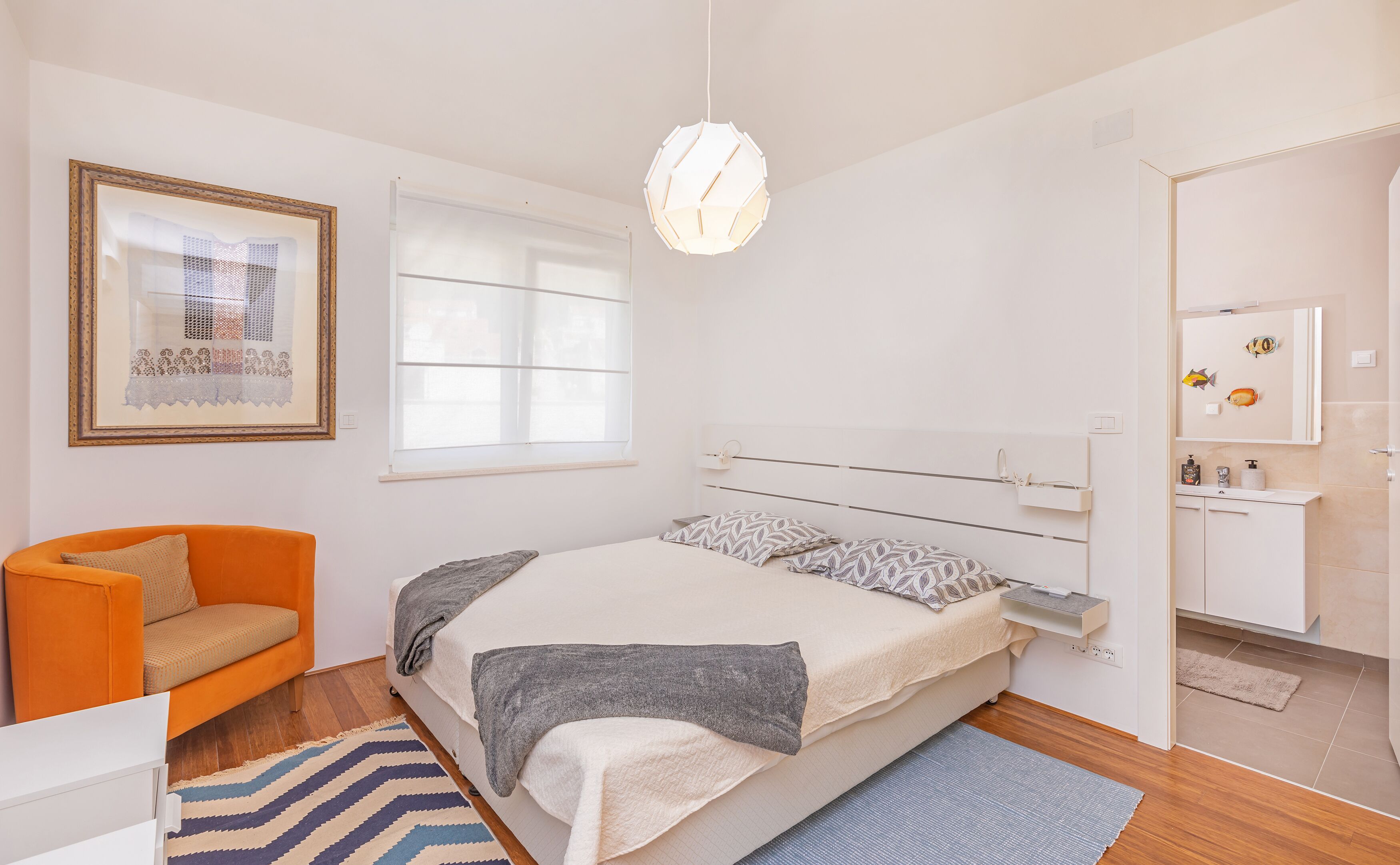 A bedroom with a bed, a chair, a bedside table, a hanging light fixture, a framed picture, and a window.