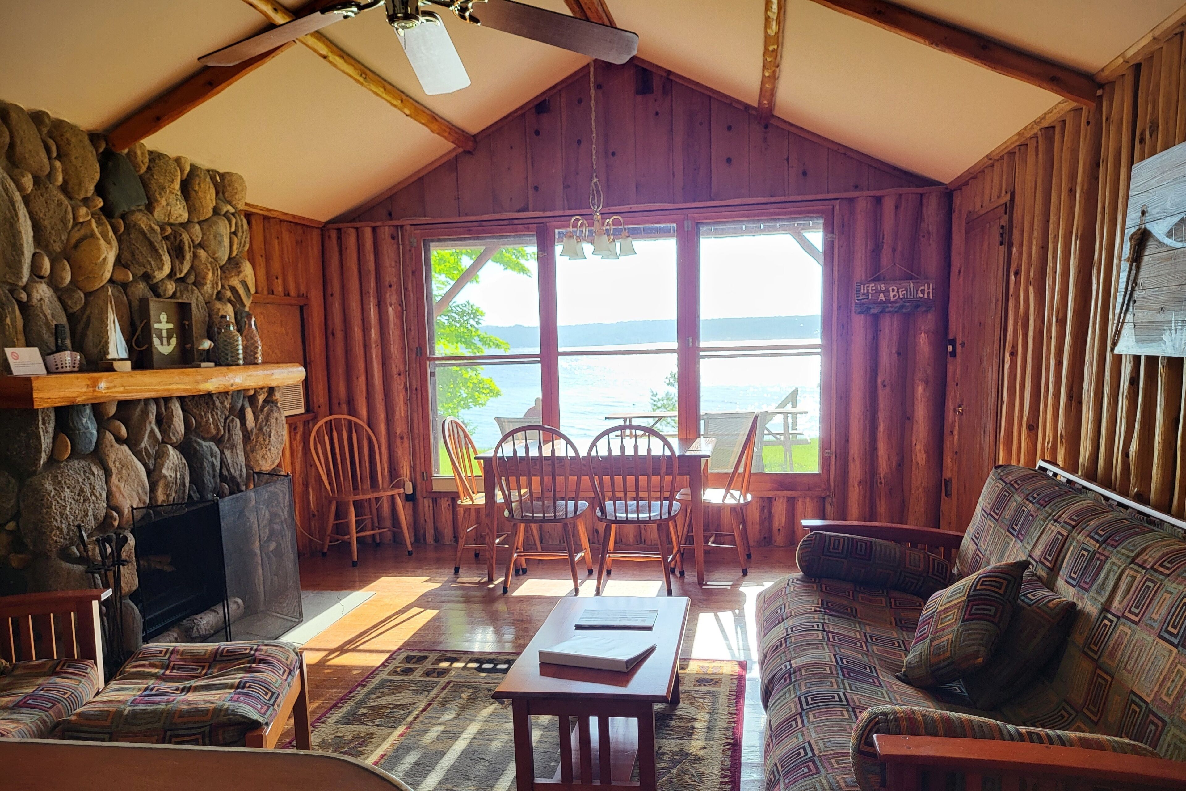 A cozy cabin with a stone wall, wooden furniture, and a large window overlooking a body of water.