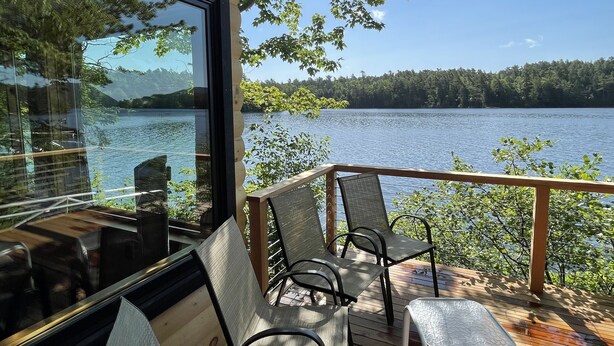 A deck with outdoor seating overlooking a lake, a glass wall, and a view of the surrounding forest.