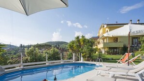 Piscina con sillas de descanso y una mesa con frutas y una botella, rodeada por una valla blanca y palmeras, con vistas a una colina y un edificio amarillo al fondo.