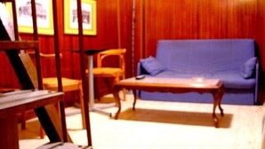 Een kamer met een blauwe bank, een houten salontafel en een stoel. Er hangen ingelijste foto's aan de muur en er staat een lamp.