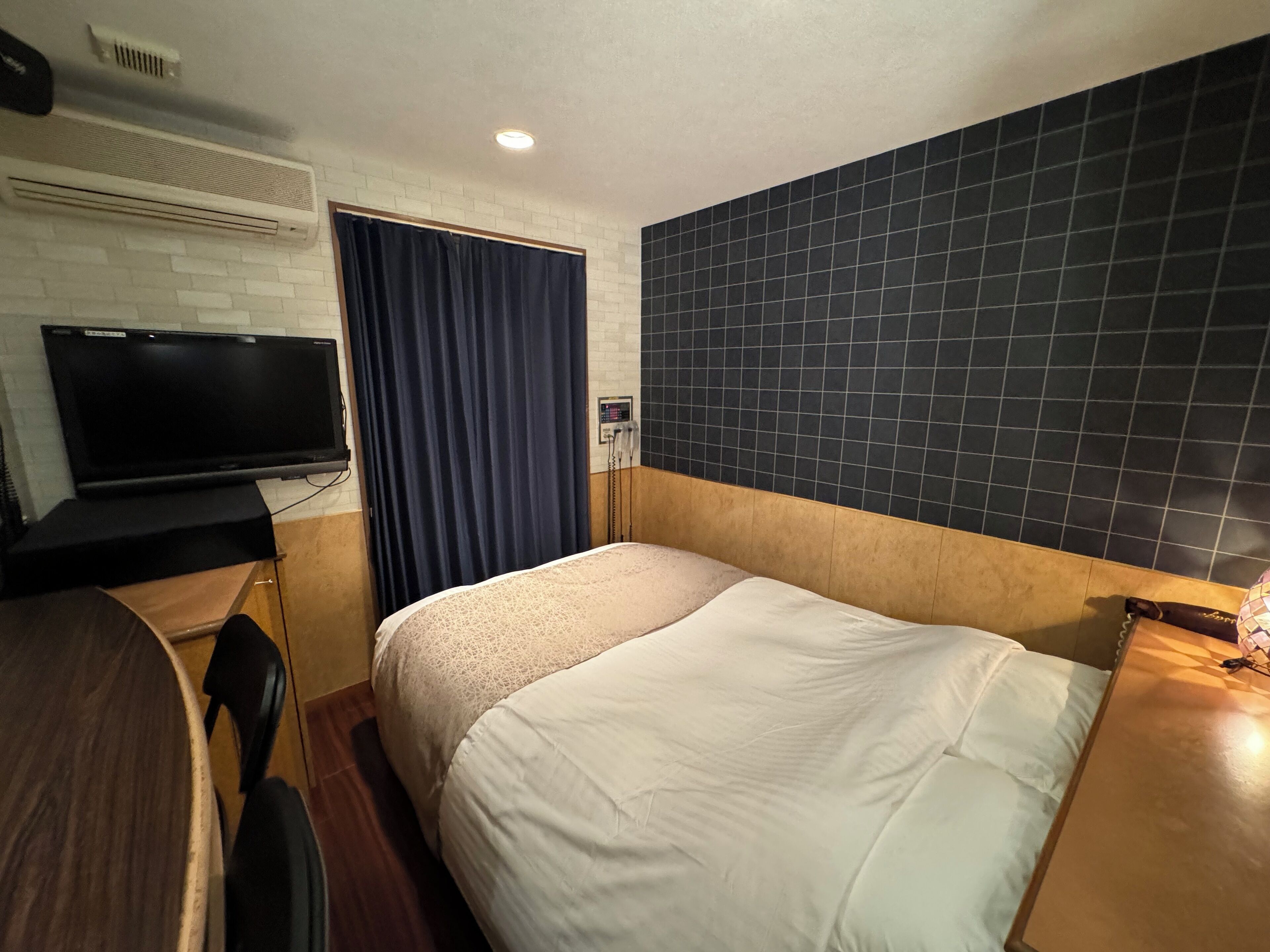 A hotel room with a bed, a desk with a flat-screen TV, a dark tiled wall, and a window with curtains.