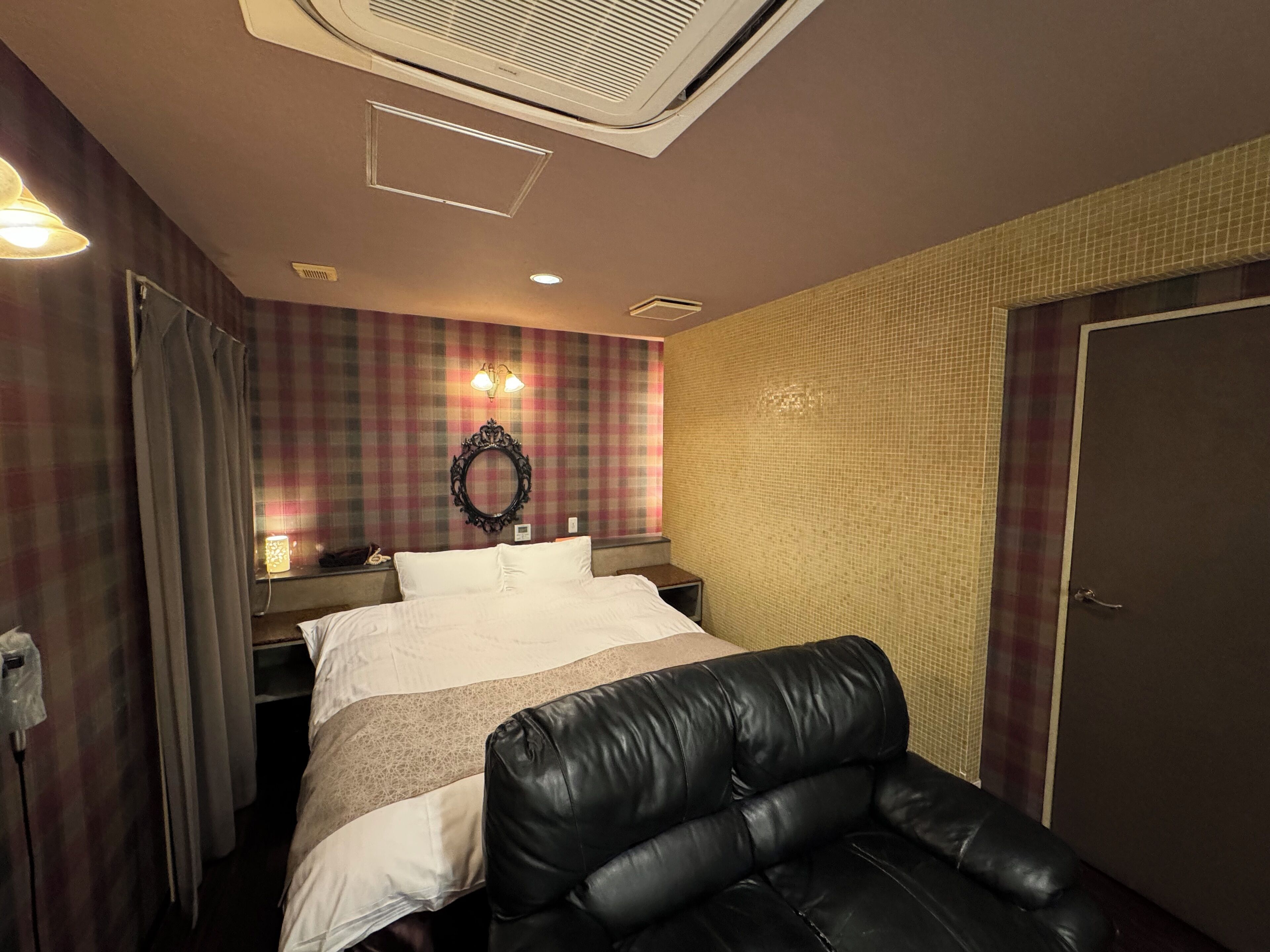 A hotel room with a bed, a black leather chair, a wall-mounted mirror, and a door.
