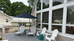 A covered deck with white chairs and a table, a large umbrella, and a view of a house with large windows.