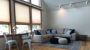 A modern living room with a beige sectional sofa, white chairs, a small table, and a large window with blinds.