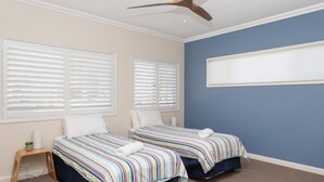 Un dormitorio con dos camas, persianas blancas, un ventilador de techo y un ventanal grande.