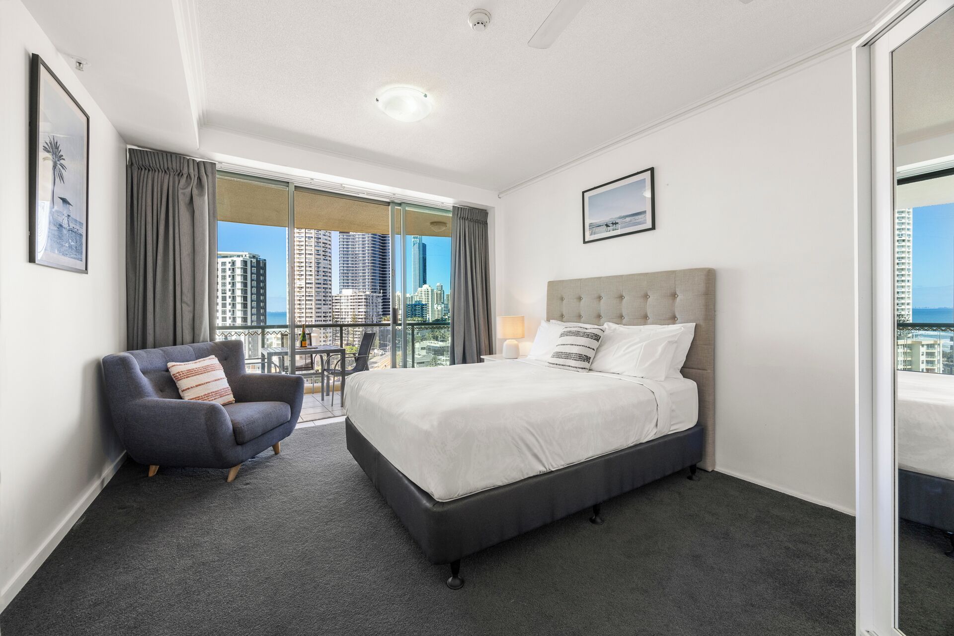 A hotel room with a bed, a chair, a balcony with a view of the city, and a framed picture on the wall.