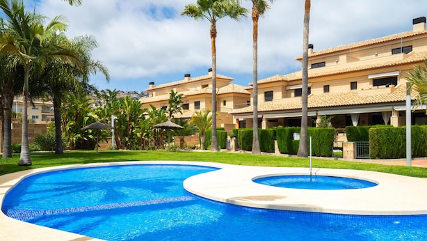 A swimming pool with a curved design, surrounded by a well-maintained lawn and palm trees. - Beautiful and cheerful holiday house in Javea, Costa Blanca, Spain  with communal pool for 2 persons (Javea)