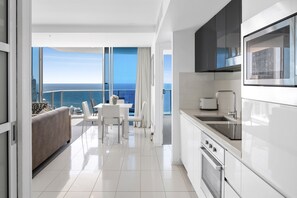 A modern kitchen with a view of the ocean, a dining area with a sofa and chairs, and a large window. - H Residences Ocean View (Surfers Paradise)