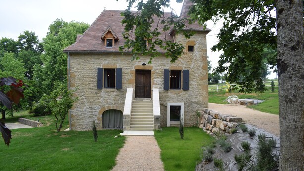 A stone house with a gravel driveway leading to a wooden door, surrounded by a well-maintained lawn and a stone wall.
