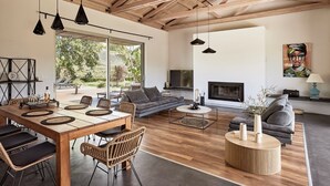 A modern living room with a wooden ceiling, a fireplace, a dining area with a long table and chairs, and a large window offering a view of the outdoors. - BBQ & Jacuzzi★HEATED pool★Big Garden★2 cars rental included★18 sleeps (Alikianos)