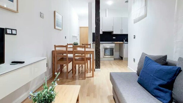 Un saló modern amb un sofà gris, una taula de dinar de fusta amb cadires, una cuina al fons i un column negre gran.