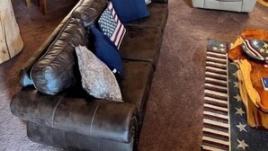Interior acogedor de una cabaña de troncos con comedor, un sofá con almohadones de la bandera estadounidense, un sillón de cuero y una pequeña mesa con alfombra a cuadros de bandera.
