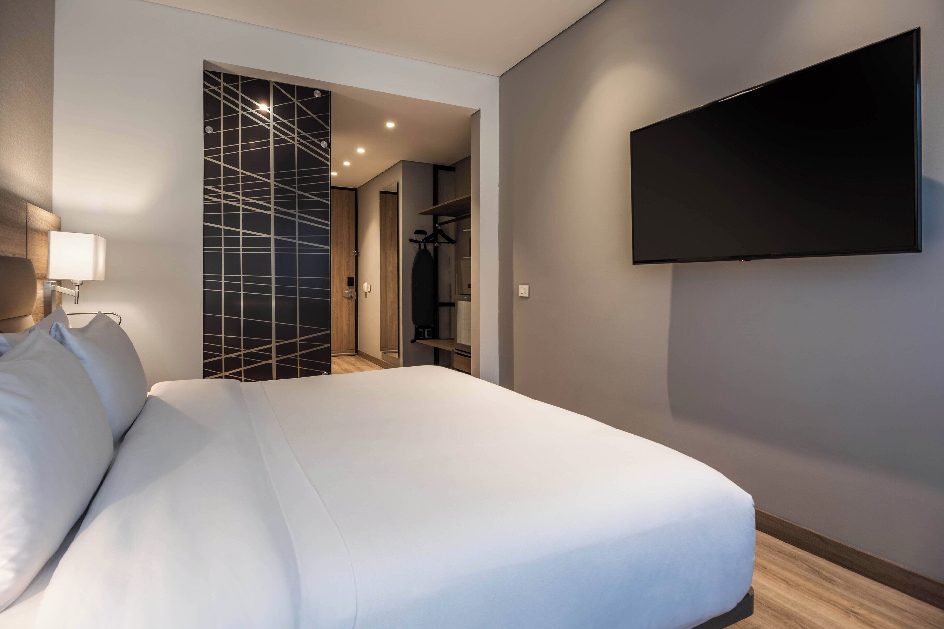 A modern hotel room with a large bed, a flat-screen TV mounted on the wall, and a walk-in closet with a mirror and shelves.