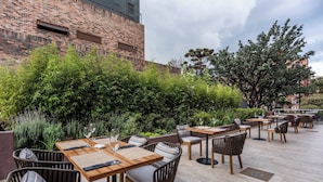 An outdoor patio with wooden tables and wicker chairs, surrounded by lush greenery and a brick wall.