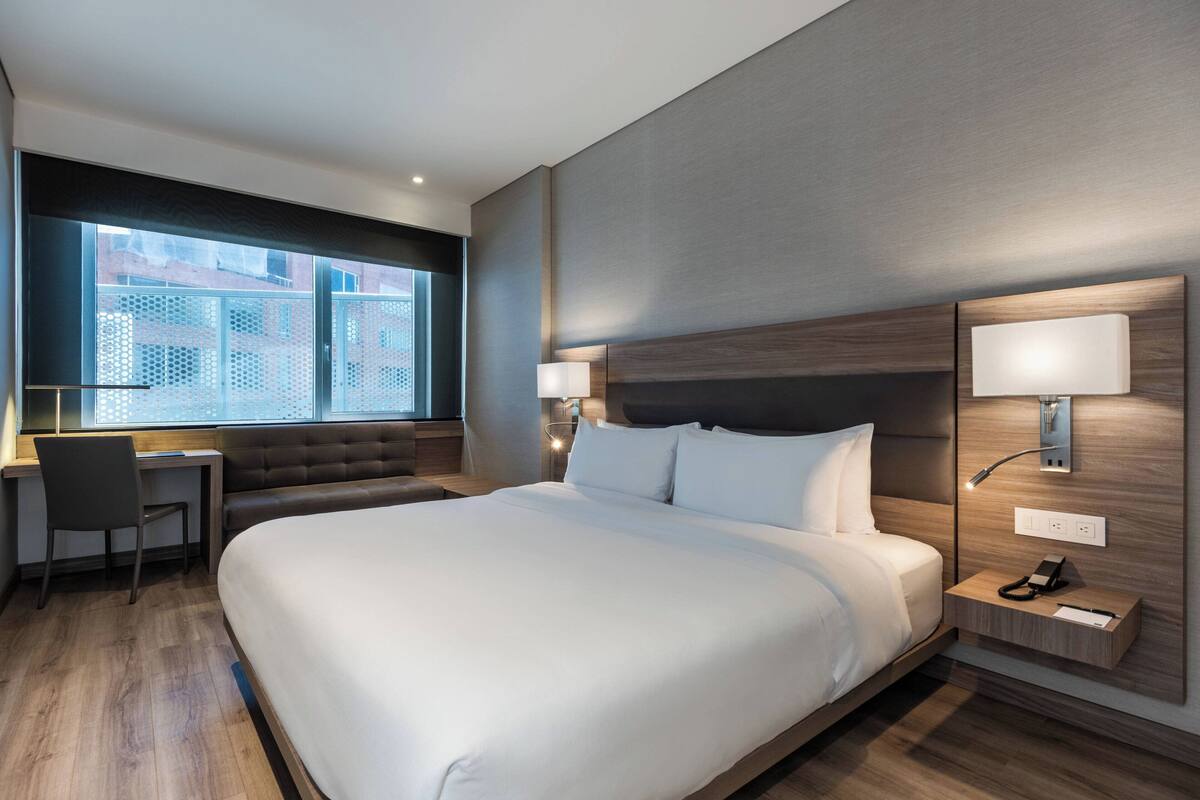 Una habitación de hotel moderna con una cama grande, un escritorio y una silla. Se ve una ventana con vistas a edificios.
