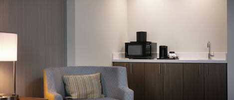 Una habitación de hotel moderna con un sillón gris, una mesita con lámpara, y una kitchenette con microondas, cafetera y lavabo.