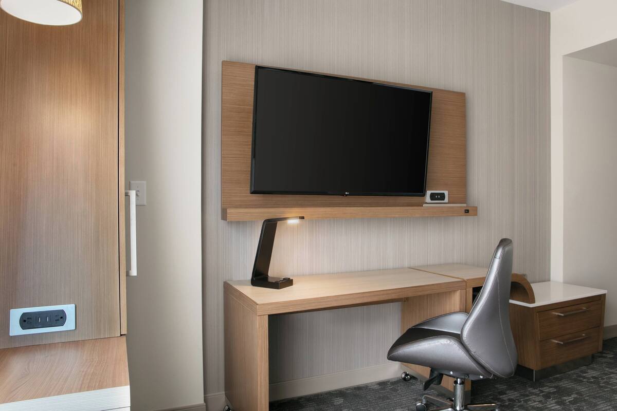 A modern hotel room with a wooden desk, a flat-screen TV mounted above it, a leather chair, and a small drawer unit.