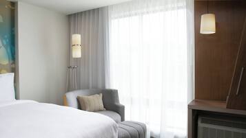 A hotel room with a bed, a grey armchair, a large window with sheer curtains, and a wooden wardrobe.
