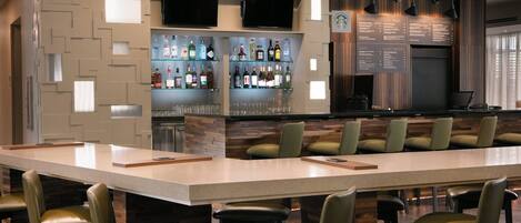 Zona de bar moderna con una barra alargada, varios taburetes y estantes llenos de botellas.