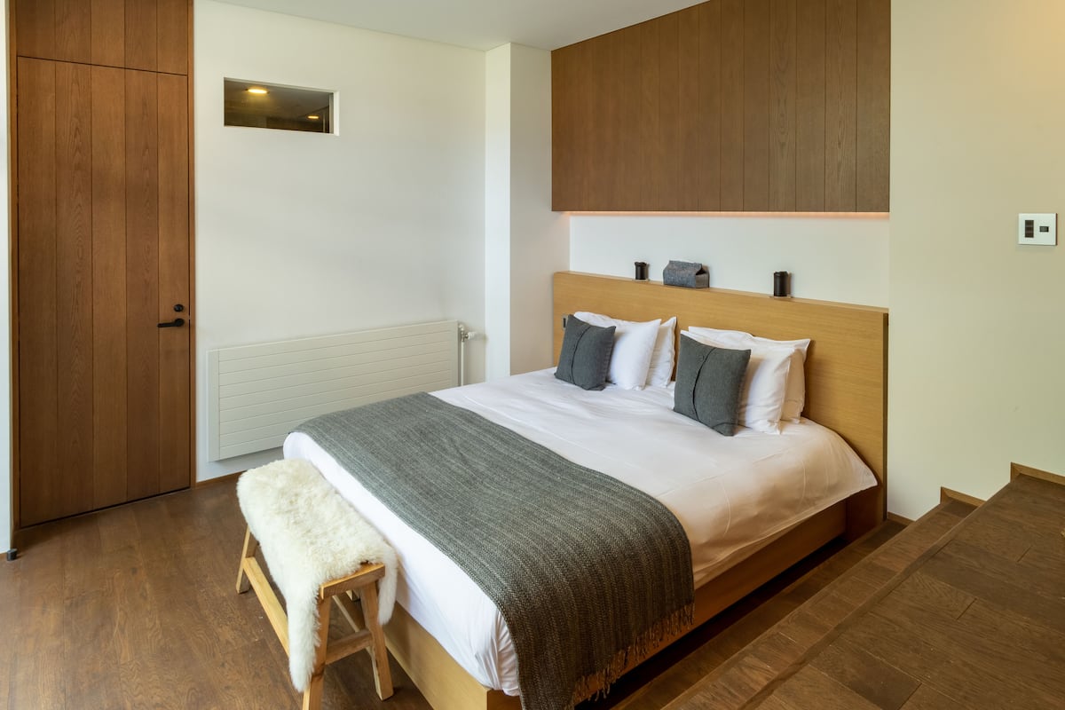 A bedroom with a wooden bed, white bedding, a grey throw blanket, and a small bedside table with a white furry throw.