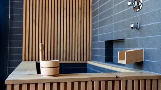 A wooden bench with a wooden bucket and a spoon, a small wooden shelf, and a wall with vertical wooden slats and a tiled section.