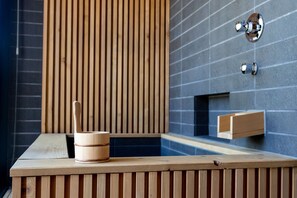 A wooden bench with a wooden bucket and a spoon, a small wooden shelf, and a wall with vertical wooden slats and a tiled section. - Yasuragi Niseko (Kutchan)
